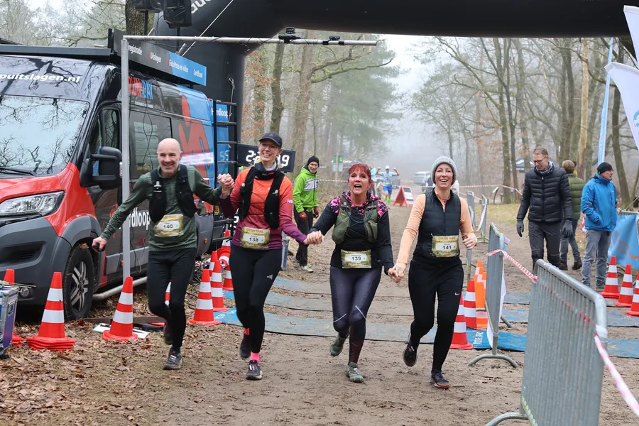 Vier lopers komen lachend en hand in hand over de finish, met publiek langs het hek