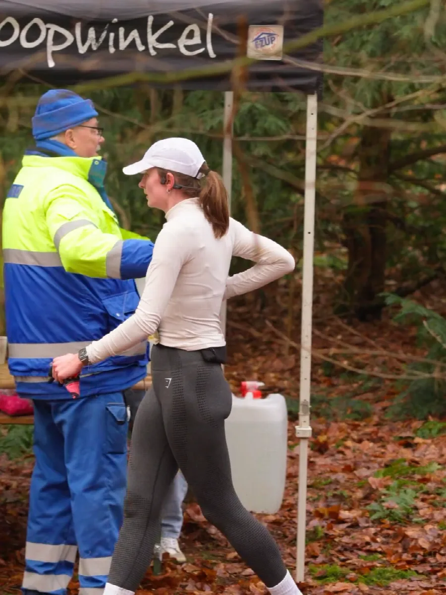Vrijwilliger in blauwe overall reikt een beker aan bij de waterpost, een loopster met witte pet pakt hem aan