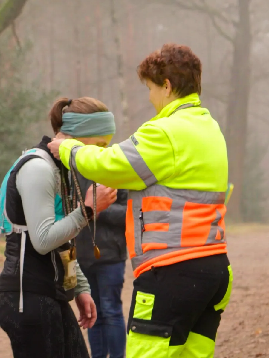 Vrijwilliger in geel hesje hangt een loopster met groene haarband een medaille om