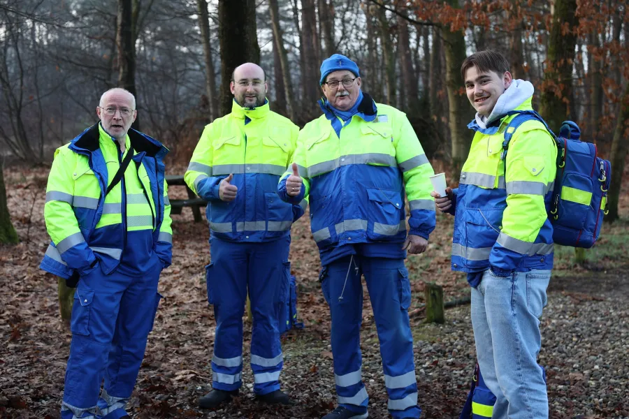 Vier EHBO-vrijwilligers in geel-blauwe jassen steken hun duim op in het bos